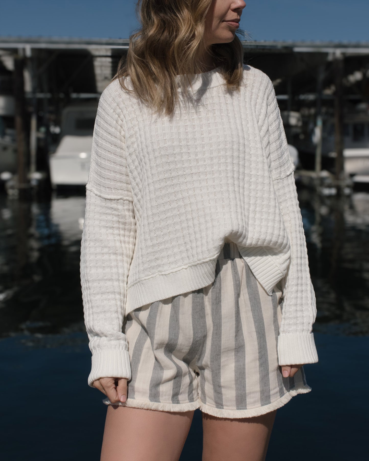 Person wearing a light sweater and striped shorts in an outdoor setting with boats in the background