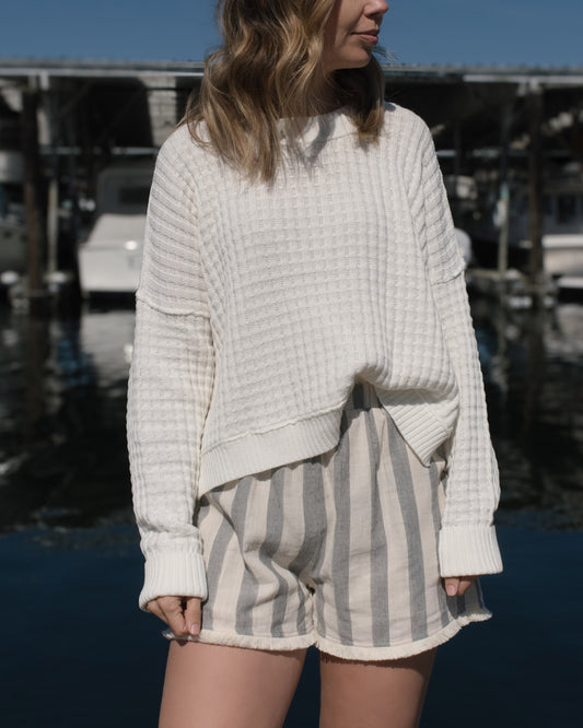 Person wearing a light sweater and striped shorts in an outdoor setting with boats in the background