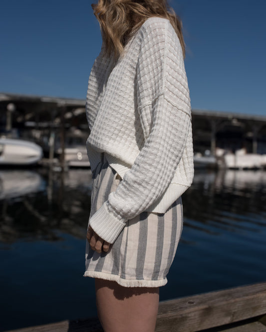Person wearing a white textured sweater and striped shorts standing on a dock with boats in the background.