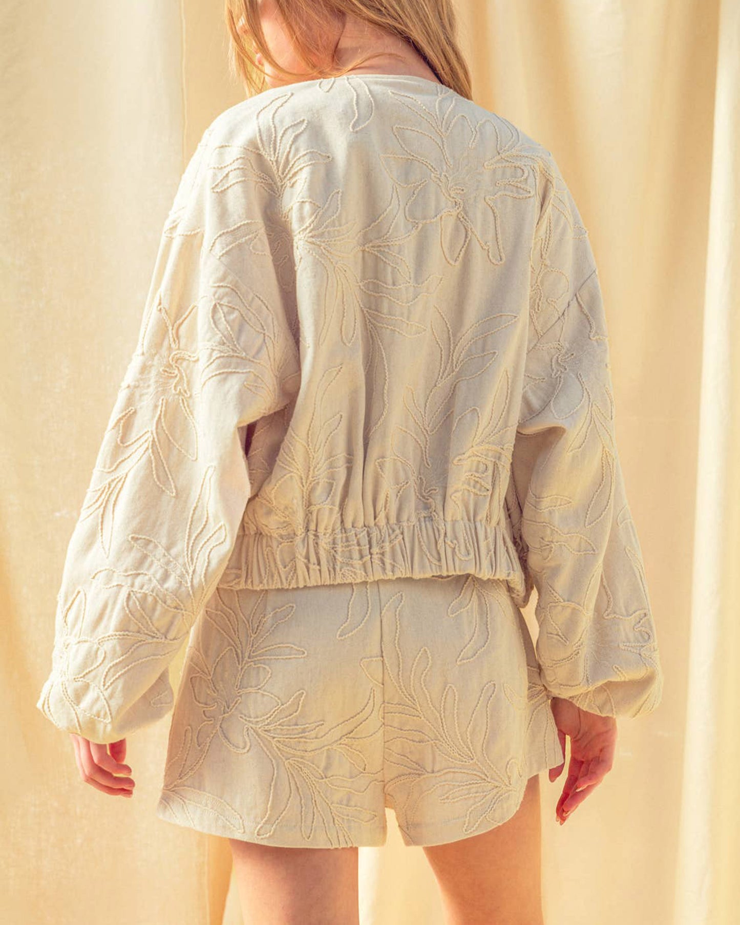 Person wearing a beige embroidered jacket and shorts set against a beige curtain.