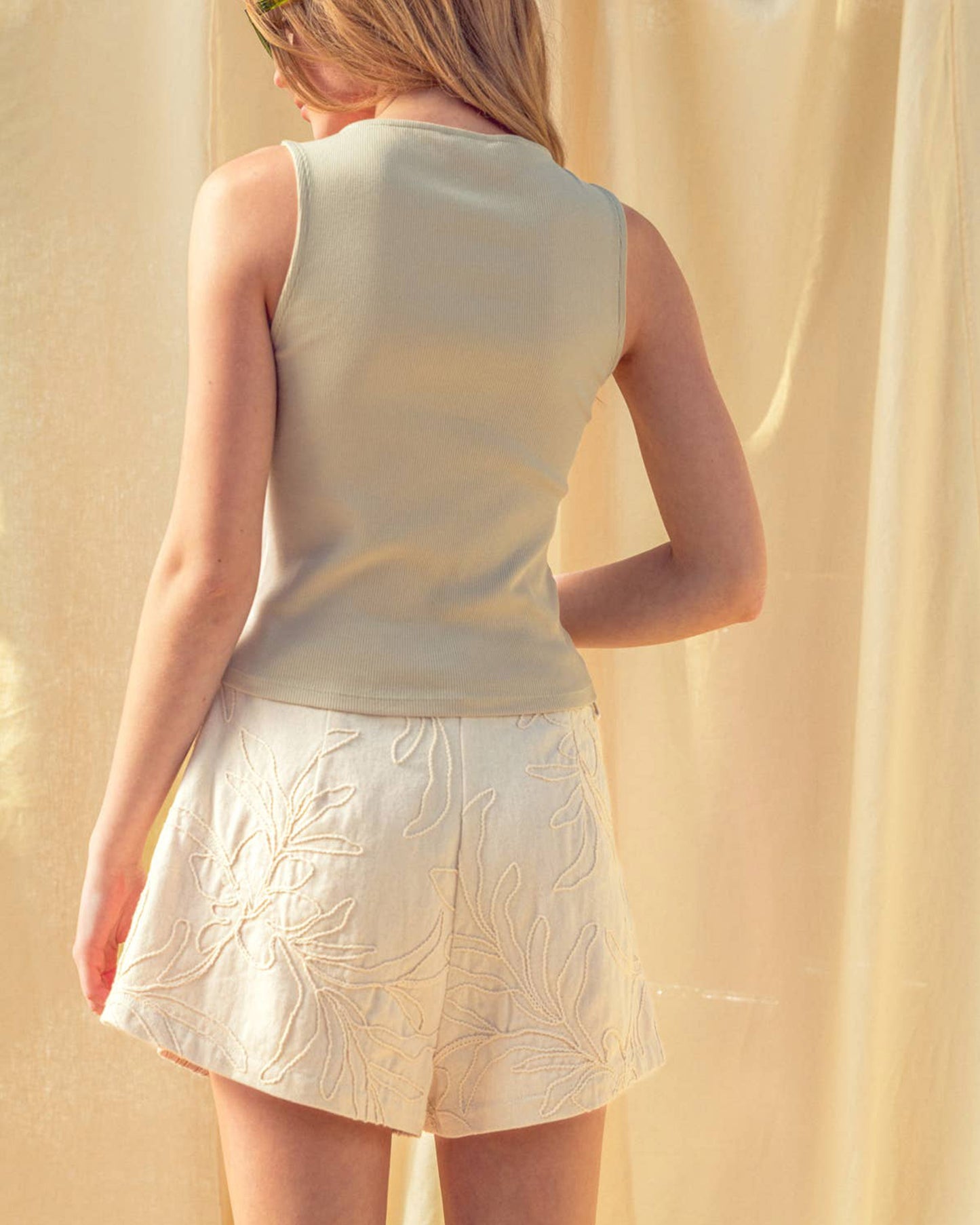 Woman wearing a light-colored sleeveless top and skirt against a beige curtain.