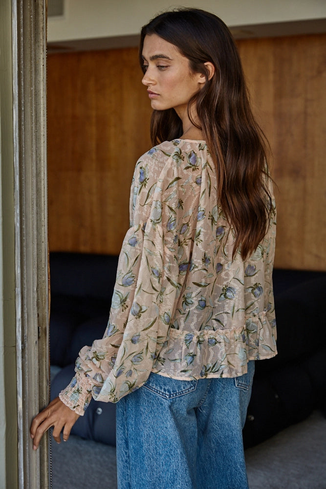 A woman wearing a beige long-sleeve blouse with a blue and green floral print, paired with blue jeans, standing in an indoor setting.

