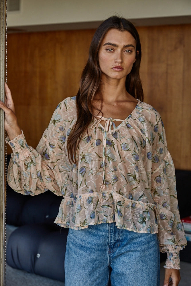 A woman wearing a beige long-sleeve blouse with a blue and green floral print, paired with blue jeans, standing in an indoor setting.