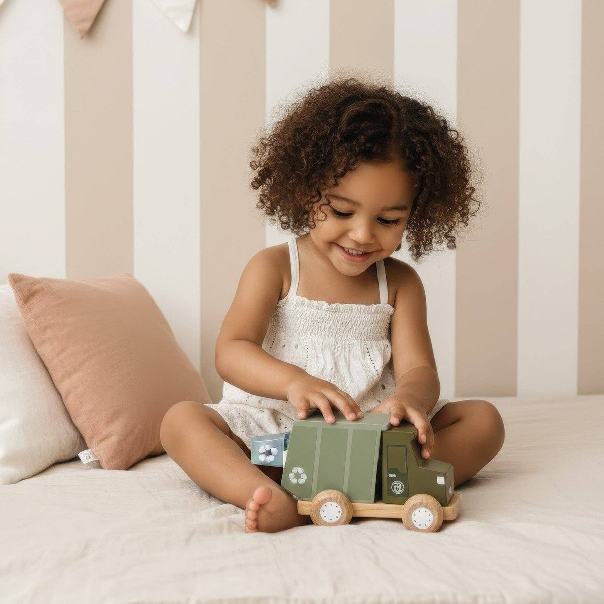 Child playing with a toy car on a bed with striped wallpaper.